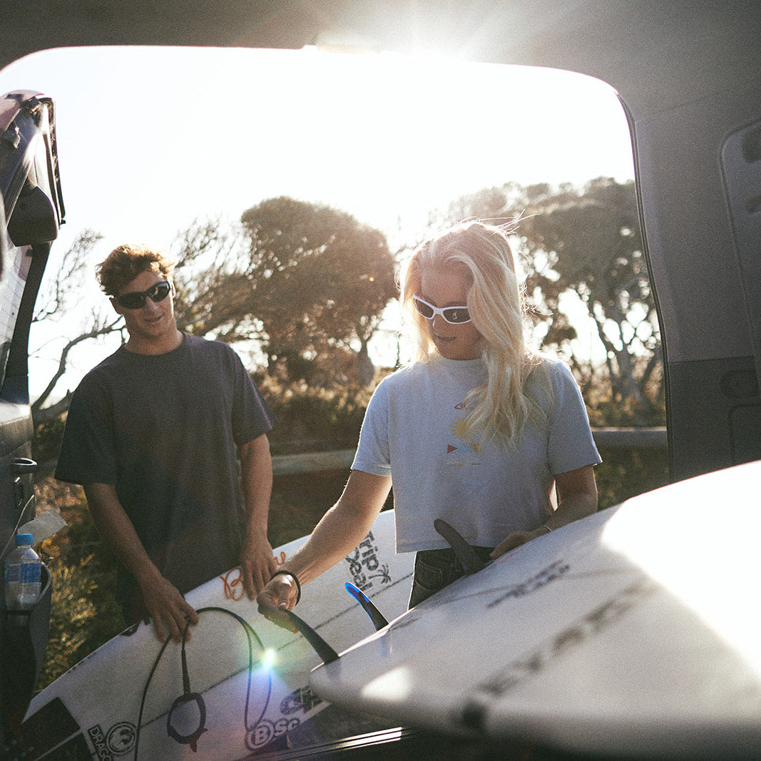 Two people wearing dragon sunglasses with surfboards standing next to a van on a sunny day.