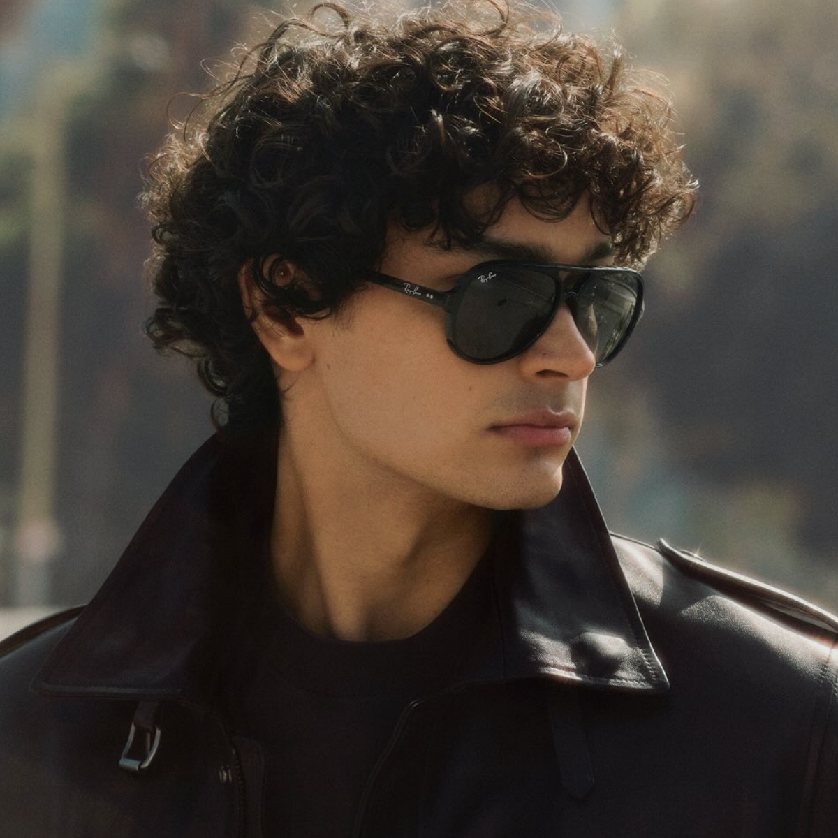 A close-up portrait of a man with curly dark hair wearing black ray-ban aviator sunglasses and a black leather jacket, 