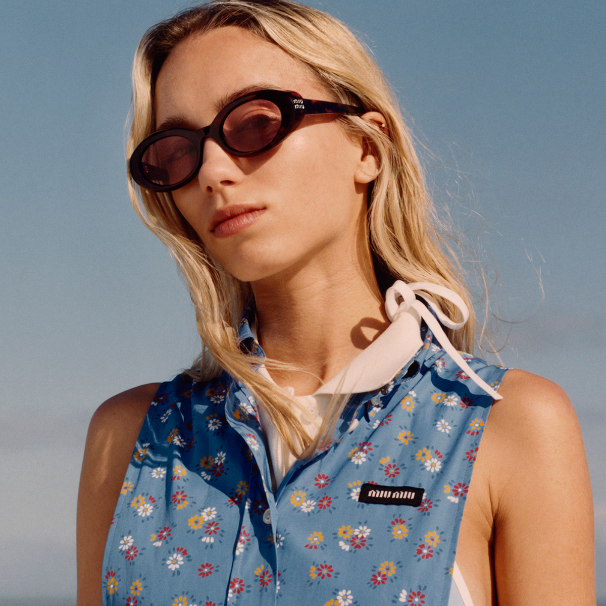 A blonde woman wearing dark brown oval sunglasses and a blue floral sleeveless top with a white collar, photographed outdoors against a clear blue sky.