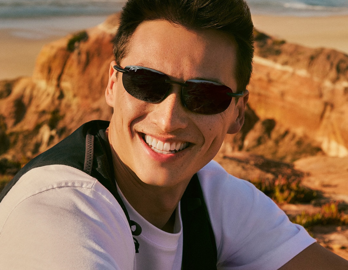 A smiling man wearing maui jim sunglasses with a white t-shirt and a black vest, photographed outdoors against a warm, rocky desert landscape.