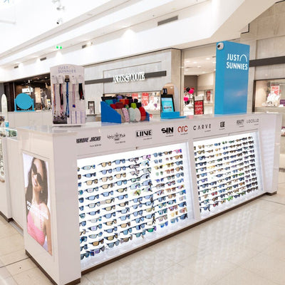 Display of sunglasses in a store with various brand names on the wall.